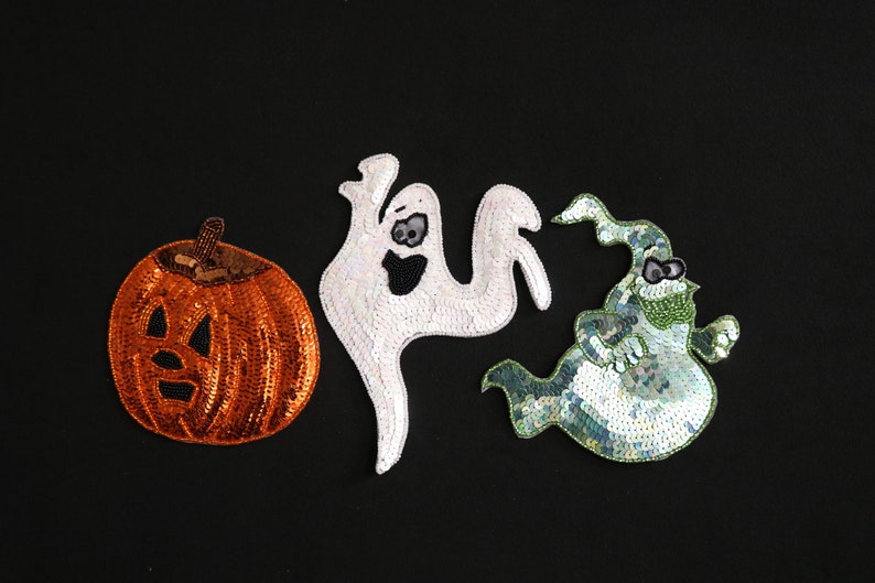 May include: Three Halloween-themed sequin patches on a black background. The patches include an orange pumpkin with a carved face, a white ghost, and a green ghost with a hat and glasses. These patches are likely for clothing or crafts.