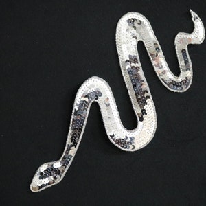 Puede incluir: Aplicación de serpiente con lentejuelas plateadas sobre fondo negro. La serpiente está en una pose sinuosa, con un patrón detallado de lentejuelas plateadas. Los bordes están delineados con hilo plateado. Esta aplicación decorativa es adecuada para ropa y accesorios.