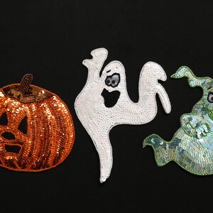 May include: Three Halloween-themed sequined decorations on a black background. The decorations include an orange pumpkin with a carved face, a white ghost, and a green ghost with a hat. The sequins give a shimmering effect.