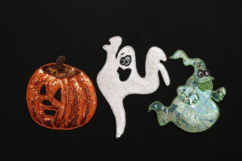 May include: Three Halloween-themed sequined decorations on a black background. The decorations include an orange pumpkin with a carved face, a white ghost, and a green ghost with a hat. The sequins give a shimmering effect.
