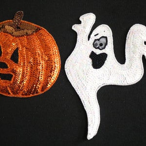 May include: Two Halloween-themed sequined patches on a black background. One is an orange pumpkin with a black beaded face and a brown stem. The other is a white ghost with a black beaded mouth and eyes.