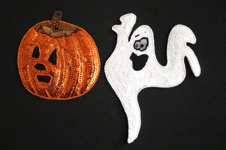 May include: Two Halloween-themed sequined patches on a black background. One is an orange pumpkin with a black beaded face and a brown stem. The other is a white ghost with a black beaded mouth and eyes.