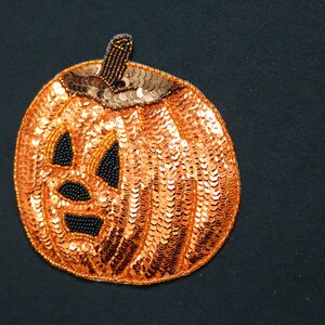 May include: An orange sequined pumpkin decoration with a smiling jack-o'-lantern face. The eyes, nose, and mouth are black beaded accents. The stem is brown with gold beads. A festive Halloween decoration.