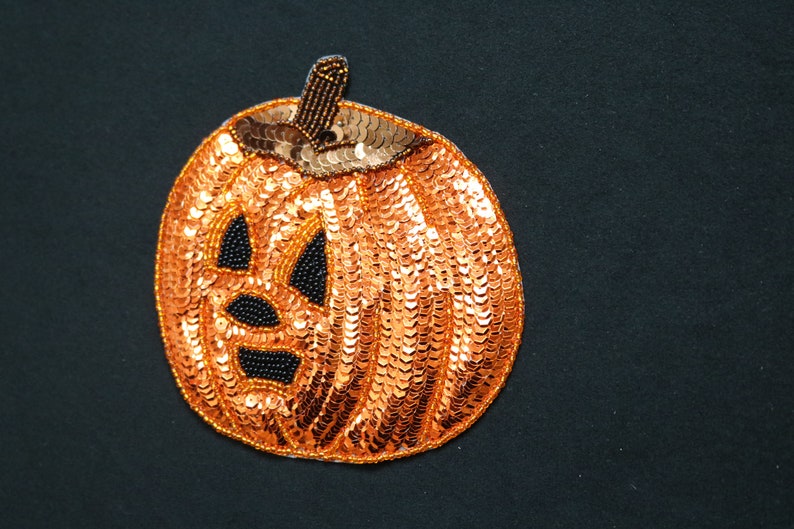 May include: An orange sequined pumpkin decoration with a smiling jack-o'-lantern face. The eyes, nose, and mouth are black beaded accents. The stem is brown with gold beads. A festive Halloween decoration.