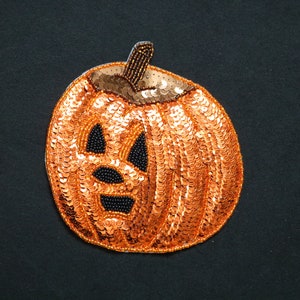 May include: A Halloween pumpkin decoration. The orange pumpkin is covered in sequins, with a carved face featuring black bead accents. The stem is brown and beaded. The background is black.
