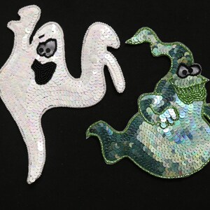 May include: Two sequinned ghost appliqu&eacute;s on a black background. One is white with black bead details for the eyes and mouth. The other is green with black and green bead details. Halloween-themed.