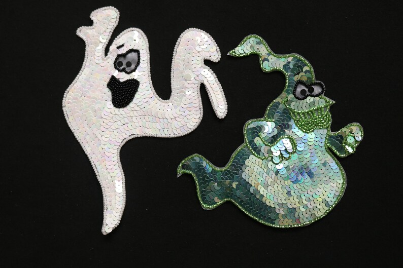 May include: Two sequinned ghost appliqu&eacute;s on a black background. One is white with black bead details for the eyes and mouth. The other is green with black and green bead details. Halloween-themed.