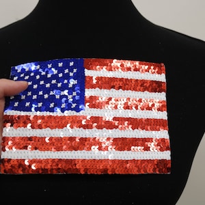 May include: A red, white, and blue American flag patch made of sequins. The flag is sewn onto a black fabric background.