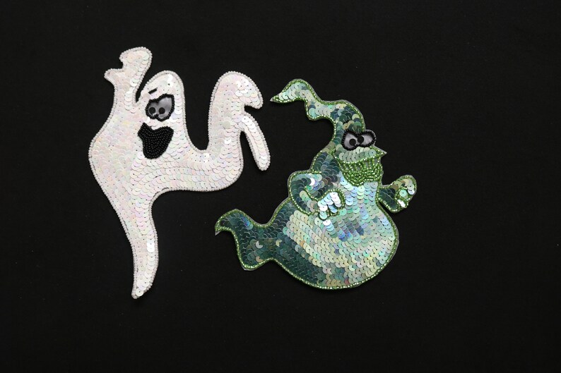 May include: Two Halloween ghost appliqu&eacute;s on a black background. One is white with black bead details, the other is green and blue with bead and sequin details. The appliqu&eacute;s are whimsical and decorative.