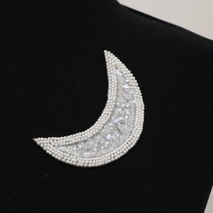 Puede incluir: Un adorno plateado en forma de media luna con brillantes transparentes y cuentas de imitación de perlas.