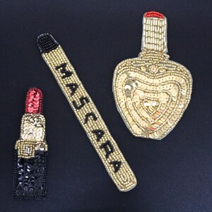 May include: Three gold and black beaded embellishments in the shape of a lipstick, mascara wand, and perfume bottle. The lipstick is red, the mascara wand says "MASCARA", and the perfume bottle is heart-shaped.