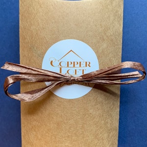 May include: A brown paper mailing envelope with a white circle sticker. The sticker has a copper colored logo that says "COPPER LOFT" with a small house icon above the text. The envelope is tied with a brown ribbon.