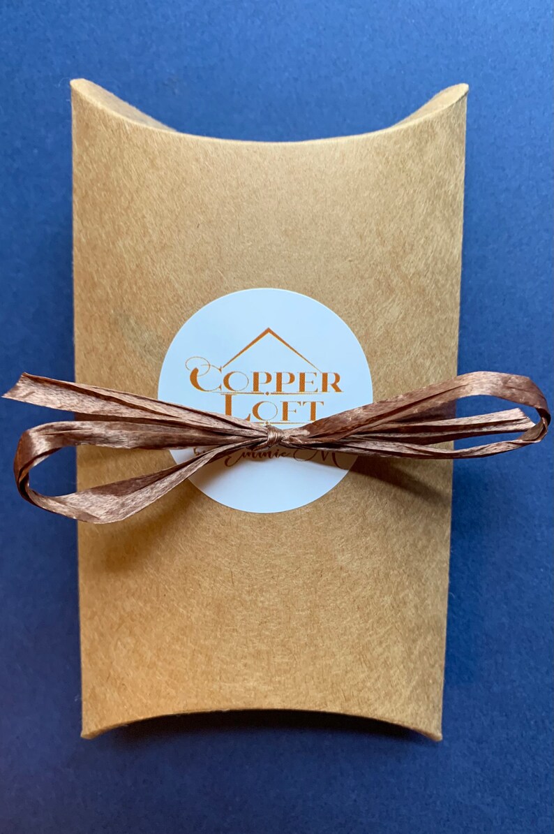 May include: A brown paper mailing envelope with a white circle sticker. The sticker has a copper colored logo that says "COPPER LOFT" with a small house icon above the text. The envelope is tied with a brown ribbon.