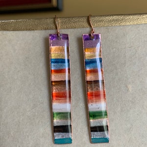 May include: A pair of long, rectangular earrings with a multi-colored striped design. The earrings are made of copper and have a silver finish. The stripes are in shades of purple, orange, red, brown, white, green, black, and blue.