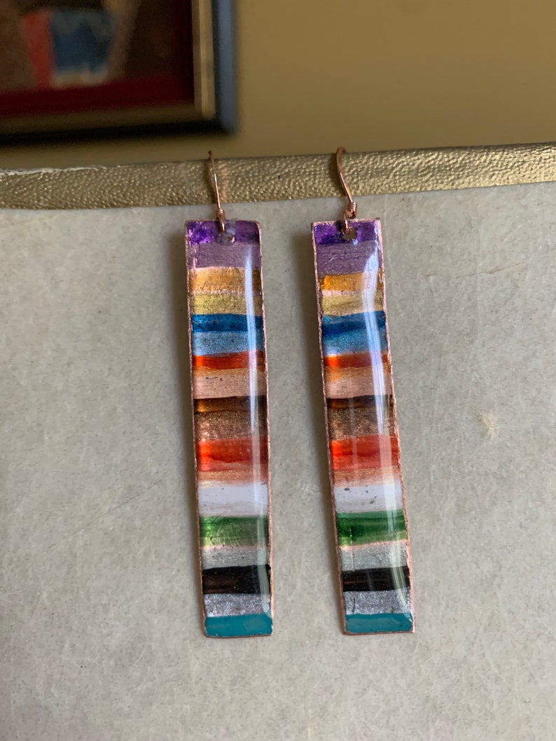 May include: A pair of long, rectangular earrings with a multi-colored striped design. The earrings are made of copper and have a silver finish. The stripes are in shades of purple, orange, red, brown, white, green, black, and blue.