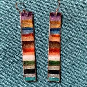 May include: A pair of long, rectangular earrings with a multi-colored striped design. The stripes are in shades of purple, gold, blue, orange, brown, white, green, black, and teal.