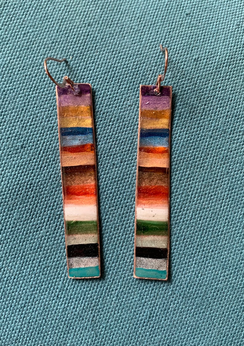 May include: A pair of long, rectangular earrings with a multi-colored striped design. The stripes are in shades of purple, gold, blue, orange, brown, white, green, black, and teal.