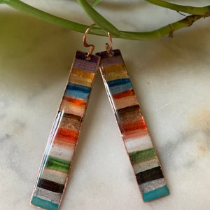 May include: A pair of long, rectangular earrings with a copper base and colorful stripes of enamel. The stripes are in shades of teal, white, black, green, orange, gold, blue, and purple.