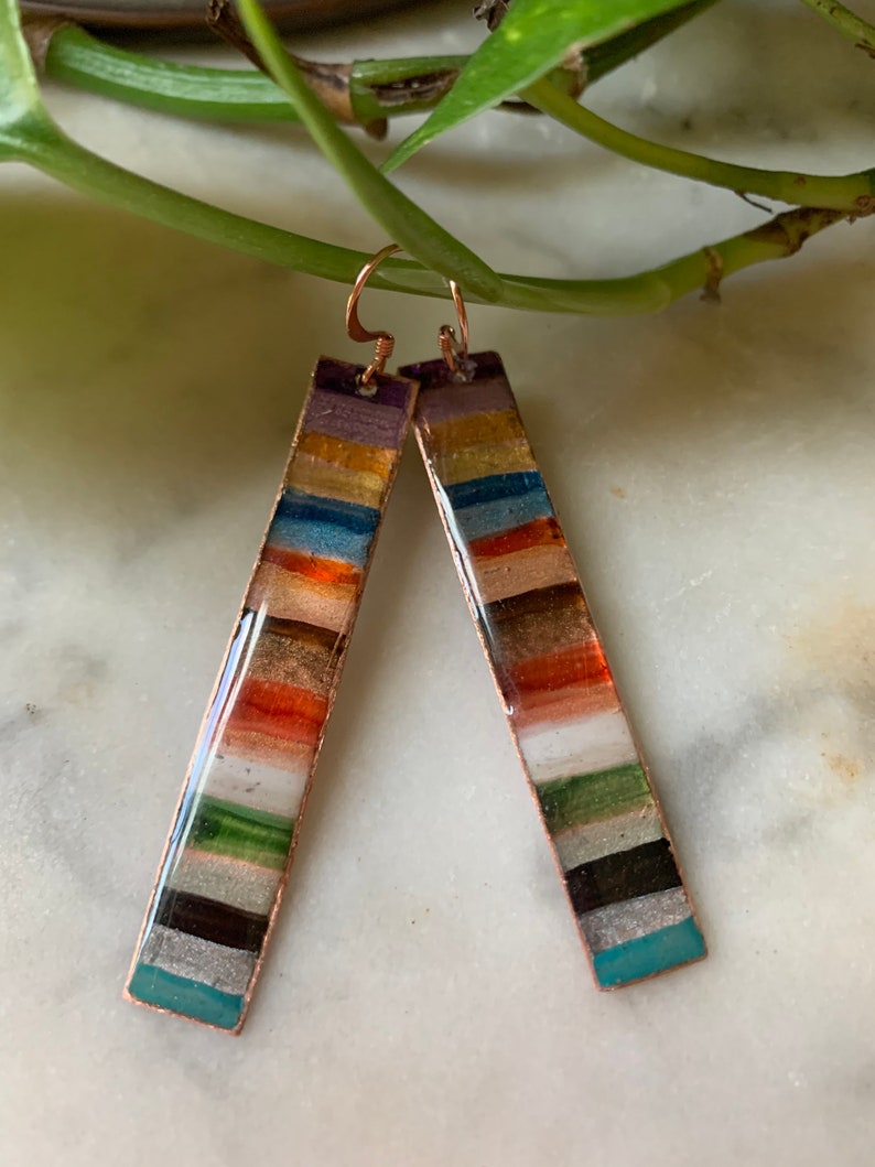 May include: A pair of long, rectangular earrings with a copper base and colorful stripes of enamel. The stripes are in shades of teal, white, black, green, orange, gold, blue, and purple.