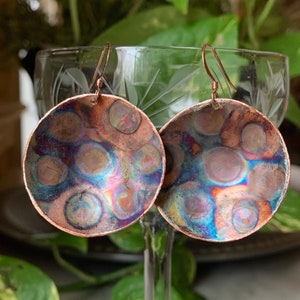 May include: A pair of copper earrings with a hammered, iridescent finish. The earrings are round and have a textured surface with a pattern of circles.