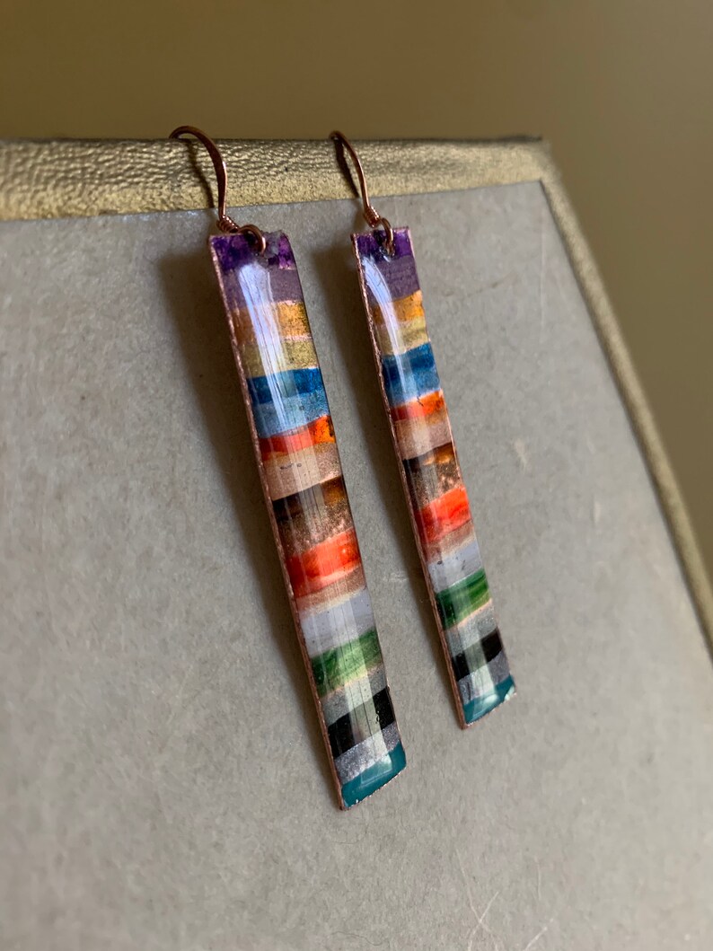 May include: A pair of long, rectangular earrings with a copper wire frame and colorful, layered, rectangular pieces of glass or stone. The earrings have a copper wire hook.