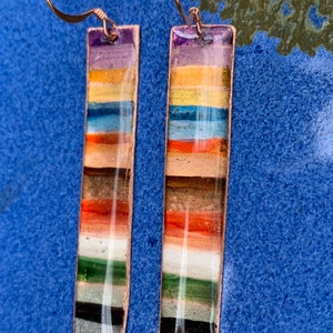 May include: A pair of long, rectangular earrings with a copper wire hook. The earrings are made of layered, colorful resin with a variety of colors, including purple, yellow, blue, orange, green, and black.