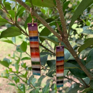 May include: A pair of colorful, striped earrings with a copper finish. Each earring is a long, thin rectangle with a variety of colors, including red, orange, yellow, green, blue, and purple.