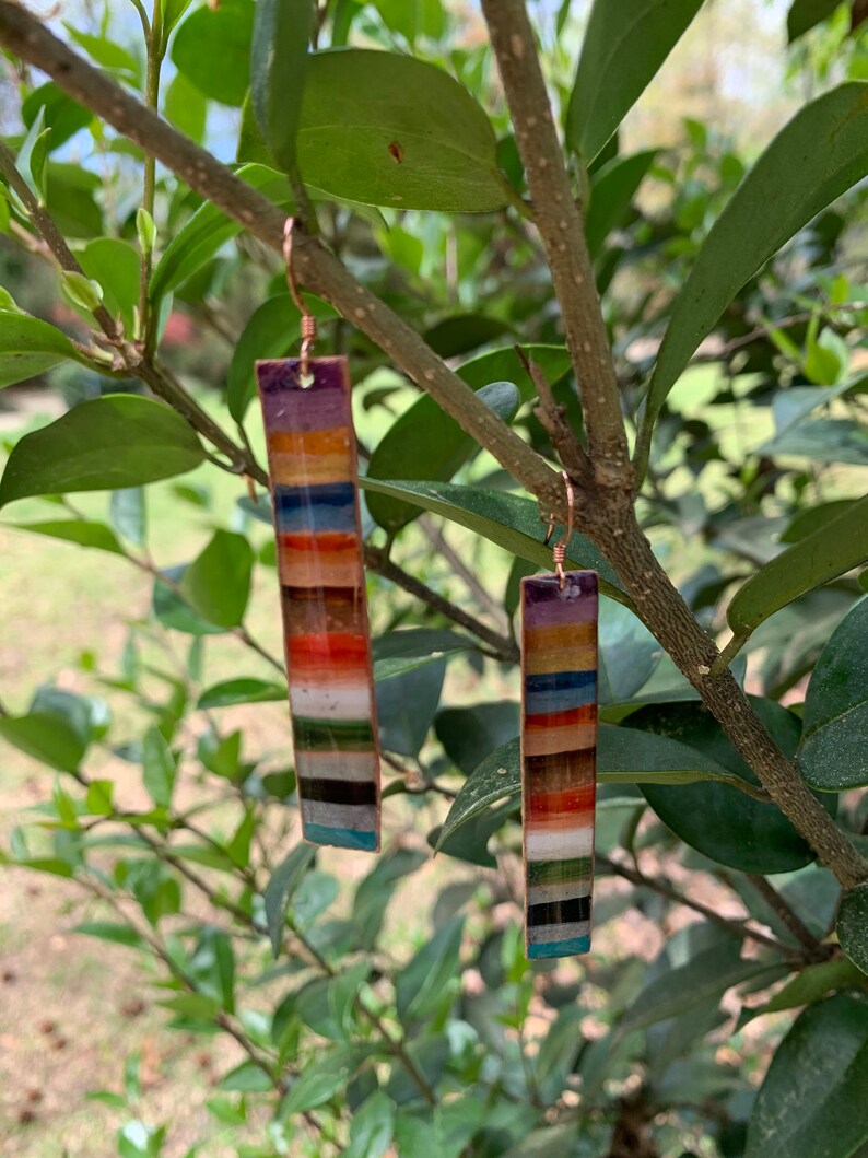 May include: A pair of colorful, striped earrings with a copper finish. Each earring is a long, thin rectangle with a variety of colors, including red, orange, yellow, green, blue, and purple.