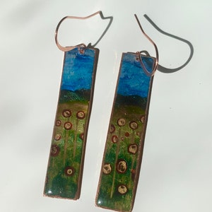 May include: A pair of dangle earrings with a copper finish. The earrings feature a rectangular design with a green, brown, and blue landscape scene. The scene includes a blue sky, green grass, and brown circles.