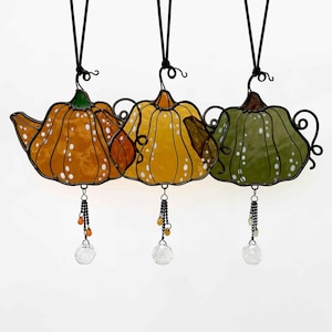May include: Three hanging stained glass pumpkin ornaments in orange, yellow, and green. Each pumpkin has white polka dots, a black metal frame, and a crystal bead at the bottom. They hang from black cords.