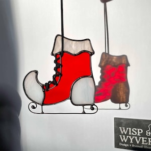May include: A stained glass Christmas ornament in the shape of a red ice skate with white accents and black laces. The ornament is hanging against a white background, with a shadow of the skate visible. The ornament is suspended by a black cord.