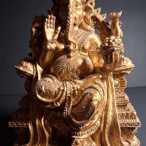 May include: A gold-colored statue of Ganesha, the Hindu god of wisdom and prosperity. The statue depicts Ganesha seated, with intricate details in the jewelry, clothing, and ornamentation. The elephant-headed deity holds a sweet treat in one hand and raises the other in a blessing gesture.