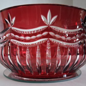 May include: A deep red glass bowl featuring intricate cut glass detailing. The upper portion displays a horizontal garland pattern with clear, etched accents, while the lower section is decorated with vertical fluted cuts around the base.