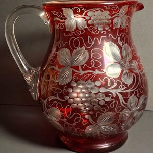 May include: A ruby red glass pitcher with a clear handle and spout. The pitcher is decorated with etched grape and leaf designs. The base is a darker shade of red. A classic piece for serving beverages.