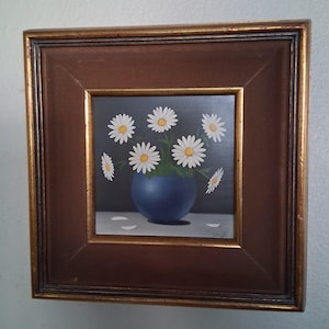 Puede incluir: Pintura enmarcada cuadrada de margaritas en un jarrón azul. Las margaritas tienen pétalos blancos y centros amarillos. El jarrón está sobre una superficie blanca contra un fondo gris oscuro. El marco es marrón con un borde interior dorado.