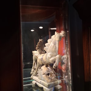 May include: A marble sculpture of a winged horse, or Pegasus, in a display case. The sculpture is a light gray color with darker gray veining. The horse is rearing up on its hind legs, with its wings spread. The sculpture is mounted on a rectangular base.