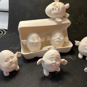 May include: Five white ceramic egg-shaped figurines with painted faces and arms. The figurines are arranged on a black surface. One figurine is standing on top of an empty egg carton.