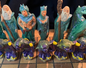 May include: A set of ceramic chess pieces with a fantasy theme. The pieces are painted in shades of green, blue, and purple with iridescent accents. The pieces include a wizard, a warrior, and a dragon.