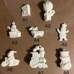 May include: A collection of eight white ceramic Christmas ornaments. The ornaments include a cheese block, hot air balloon, Santa Claus on a chimney, a sleigh, a snowman, Santa on a motorcycle, a train, and a snowman with a top hat.
