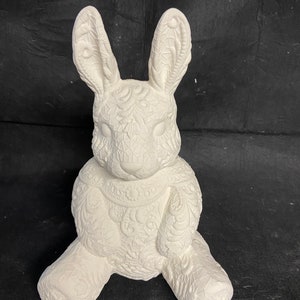 May include: A white ceramic bunny figurine with detailed floral and geometric patterns. The bunny is sitting with its legs extended and ears upright. The figurine is unpainted, ready for customization.