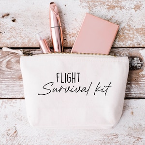 Könnte beinhalten: Eine weiße Canvas-Reißverschlusstasche mit dem Text "FLIGHT Survival Kit" in schwarzen Buchstaben. Die Tasche ist gefüllt mit Make-up-Artikeln, darunter eine roségoldene Kompaktdose und eine schwarze Mascara-Tube.