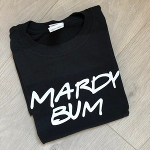 May include: Black t-shirt with white text that says "Mardy Bum".