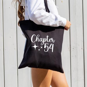May include: Black tote bag featuring the text "Chapter 54" in white script with star details. The bag is slung over a shoulder, suggesting it's made from durable material. A light-coloured wooden wall forms the backdrop.