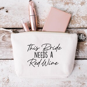 May include: White canvas makeup bag with a zipper closure. The bag has black text that reads "This Bride Needs a Red Wine".