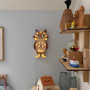 May include: A wooden robot-shaped wall light with warm LED lights is mounted on a light blue wall. A wooden house, a blue speaker, and other wooden toys sit on a wooden table. A backpack hangs on the wall to the left.