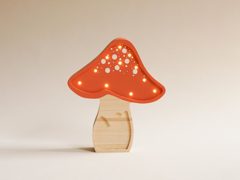 May include: A mushroom-shaped lamp featuring a light orange cap with white circular cutouts and integrated warm LED lights. The base is constructed from light-colored wood with a visible grain, providing a natural contrast to the orange top.
