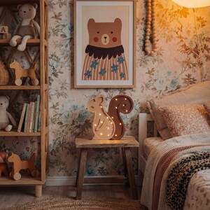 May include: A children's bedroom scene with a wooden bookshelf, a framed bear illustration, and a wooden squirrel-shaped night light. The room has floral wallpaper, a round jute rug, and a bed with a patterned blanket.