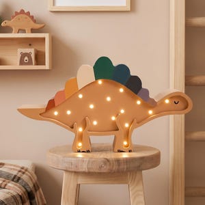 May include: A wooden dinosaur-shaped night light with a warm, amber glow. The stegosaurus has a colorful spine and is lit with small bulbs. It sits on a wooden stool, with a framed print and other decor in the background.