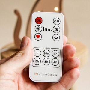 May include: A white remote control with black buttons and icons, including "ON," "OFF," a sun, a heart, and a moon. The remote also has timer settings for 30m, 1h, 2h, 3h, and 4h. The brand name "roomGAGA" is printed at the bottom.