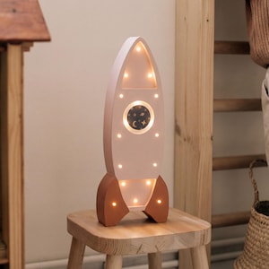 Op de afbeelding: Een houten nachtlampje in de vorm van een raket, in zachtroze en bruin. De raket heeft een rond raam met een maan- en sterrenmotief en verschillende kleine lampjes. Het staat op een kleine houten kruk.